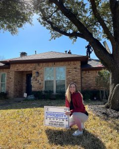 A woman poses next to a Priority Roofing Yard sign